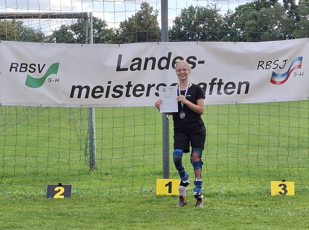Eine junge Sportlerin mit Beinorthesen hält Medaille und Urkunde auf Platz 1 vor dem Banner der Landesmeisterschaften.