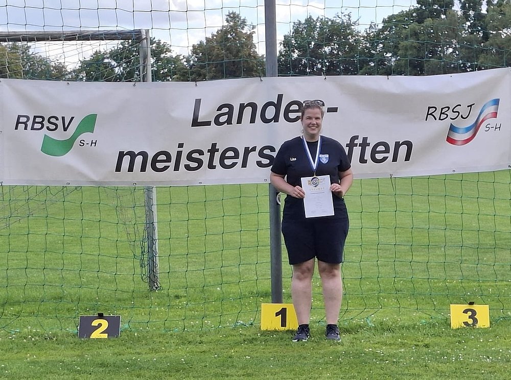 Eine Sportlerin in dunkler Sportkleidung mit Medaille und Urkunde auf Platz 1 vor dem Banner der Landesmeisterschaften.