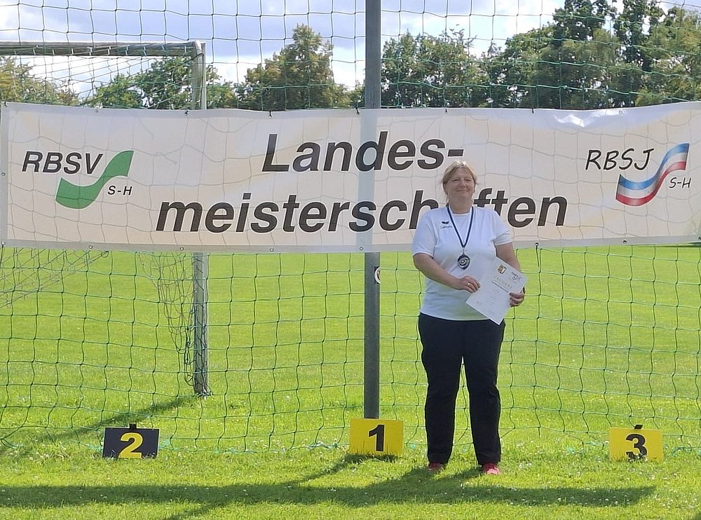 Eine Sportlerin in weißem Shirt mit Medaille und Urkunde auf Platz 1 vor dem Banner der Landesmeisterschaften.