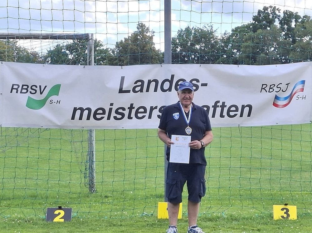 Ein Sportler mit Basecap, Medaille und Urkunde auf Platz 1 vor dem Banner der Landesmeisterschaften.