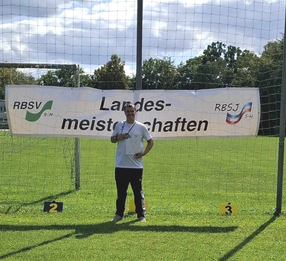 Ein Sportler in weißem Shirt mit Medaille und Urkunde vor dem Banner der Landesmeisterschaften.