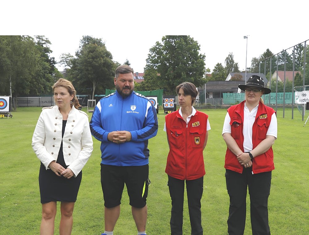 Begrüßung auf dem Sportplatz: eine Rednerin, der Ausrichter in blauer Vereinsjacke und zwei Kampfrichterinnen in roten Westen, im Hintergrund die Zielscheiben.