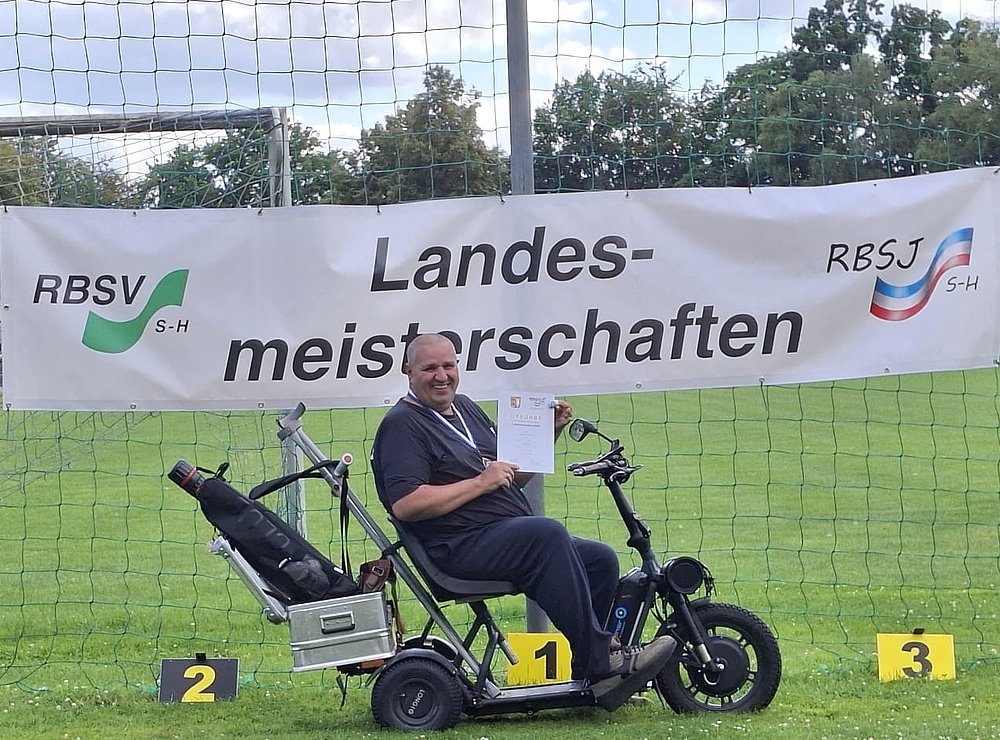 Ein Sportler auf einem Elektromobil hält lächelnd seine Urkunde in die Kamera, daneben das Schild für Platz 1.