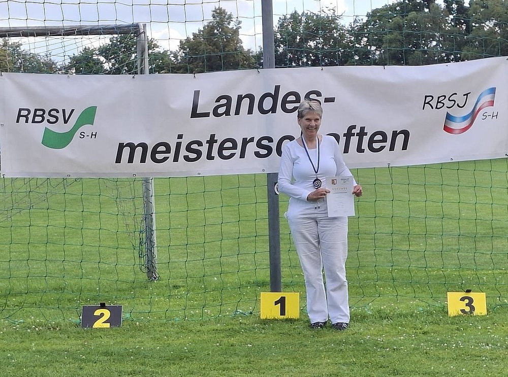Eine Sportlerin in weißer Sportkleidung mit Medaille und Urkunde auf Platz 1 vor dem Banner der Landesmeisterschaften.