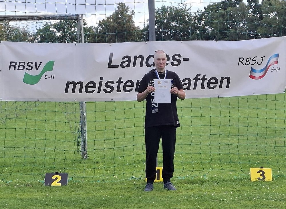 Ein Sportler in schwarzem Shirt mit Medaille und Urkunde auf Platz 1 vor dem Banner der Landesmeisterschaften.