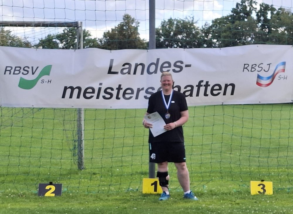 Eine Sportlerin mit Medaille und Urkunde auf Platz 1 vor dem Banner der Landesmeisterschaften.